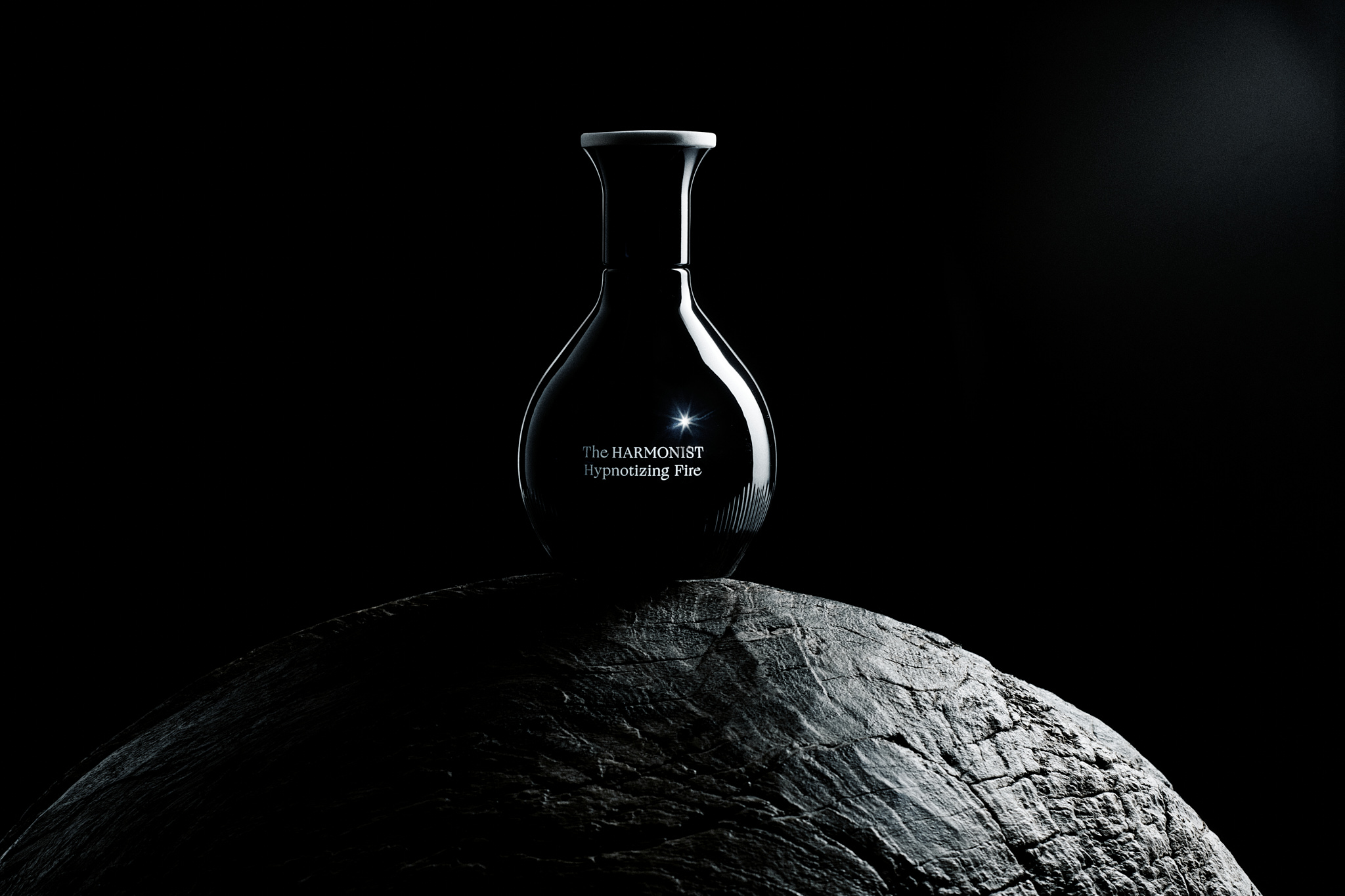 Dark reflective perfume bottle labeled The HARMONIST Hypnotizing Fire on textured stone sphere.