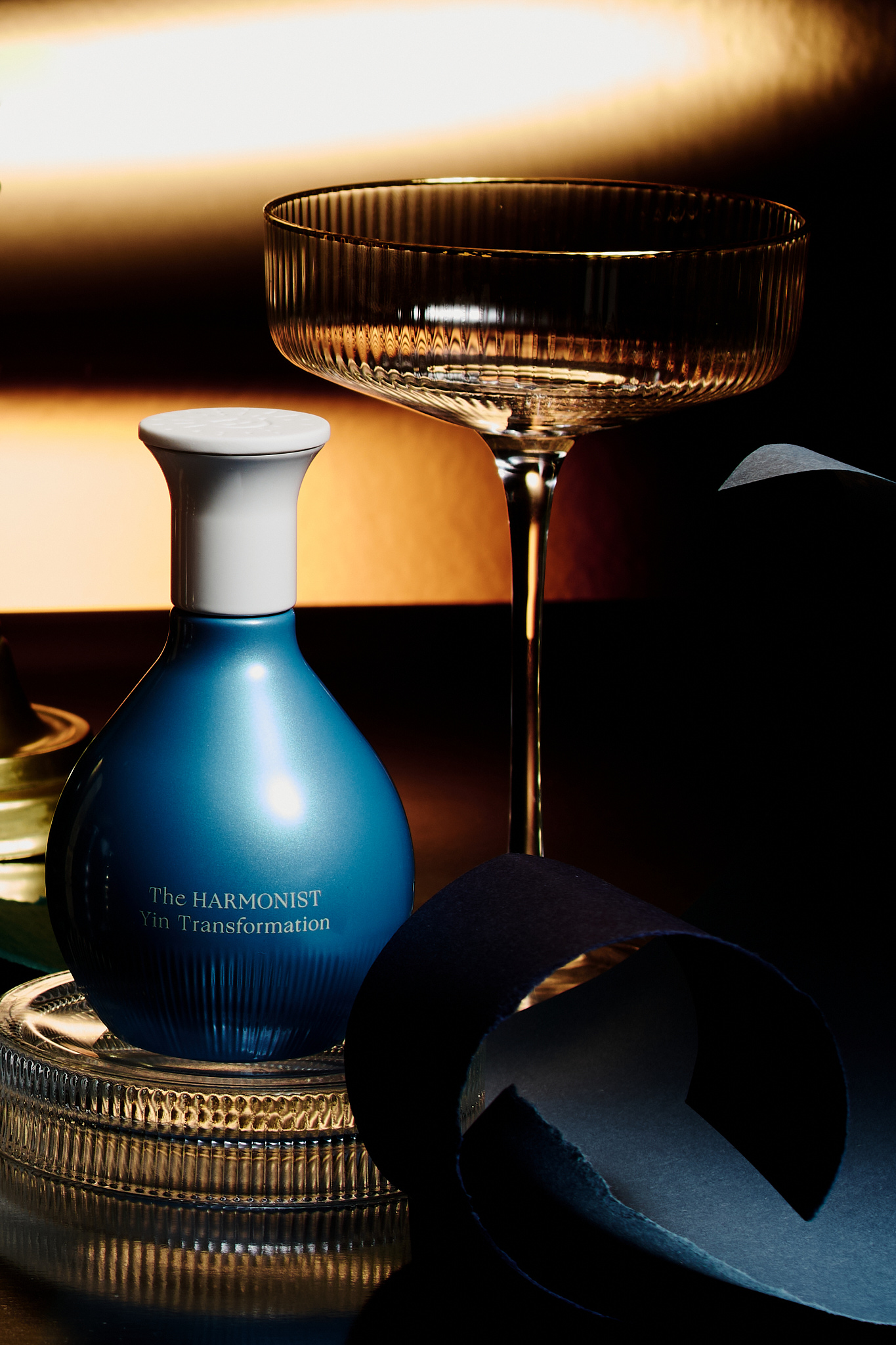 Blue bottle labeled The Harmonist Yin Transformation on textured glass plate beside empty ribbed stemmed glass and curled dark paper.