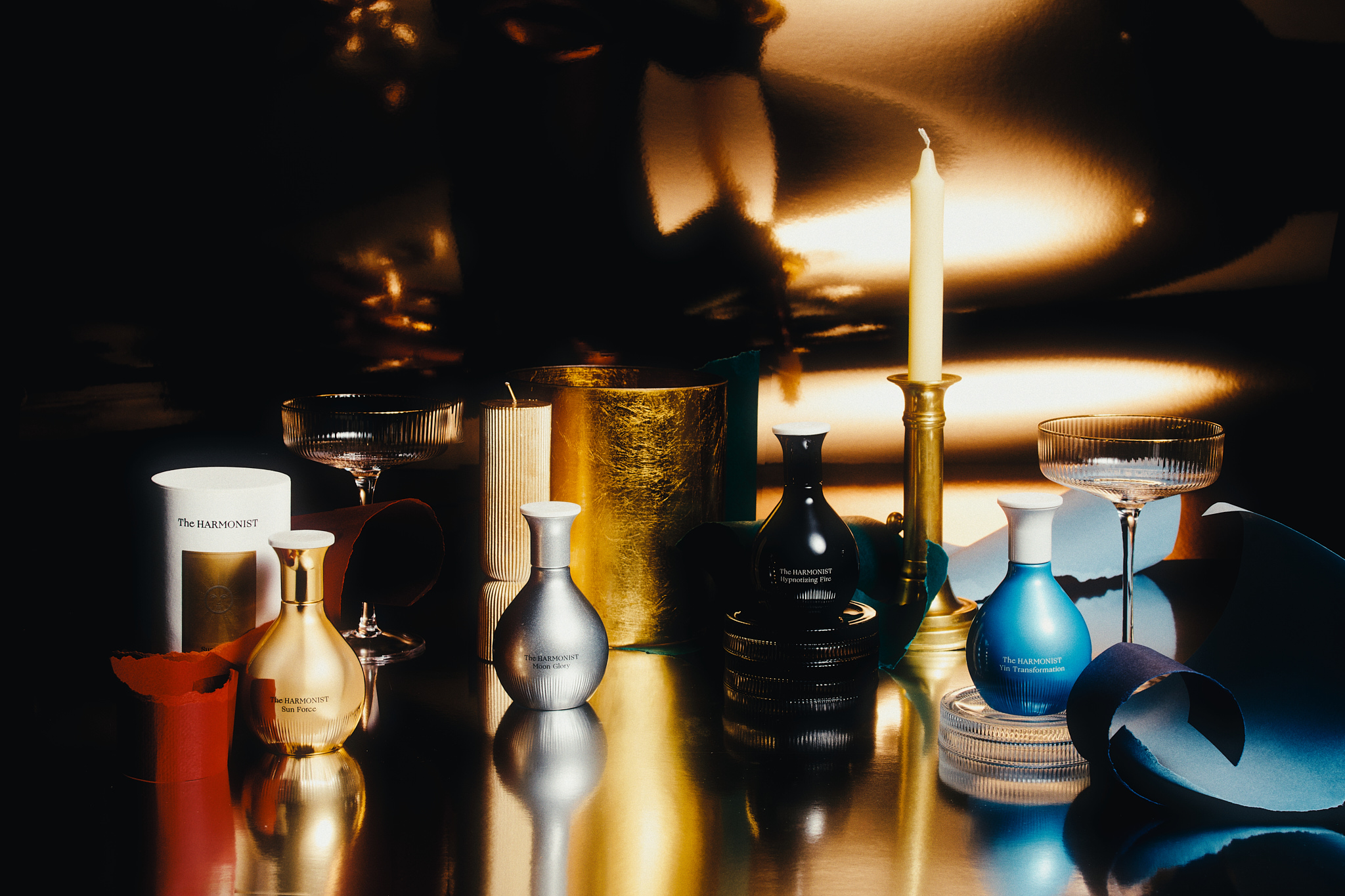 Four colored bottles labeled The Harmonist with candles, glassware, and decorative paper on a reflective surface.