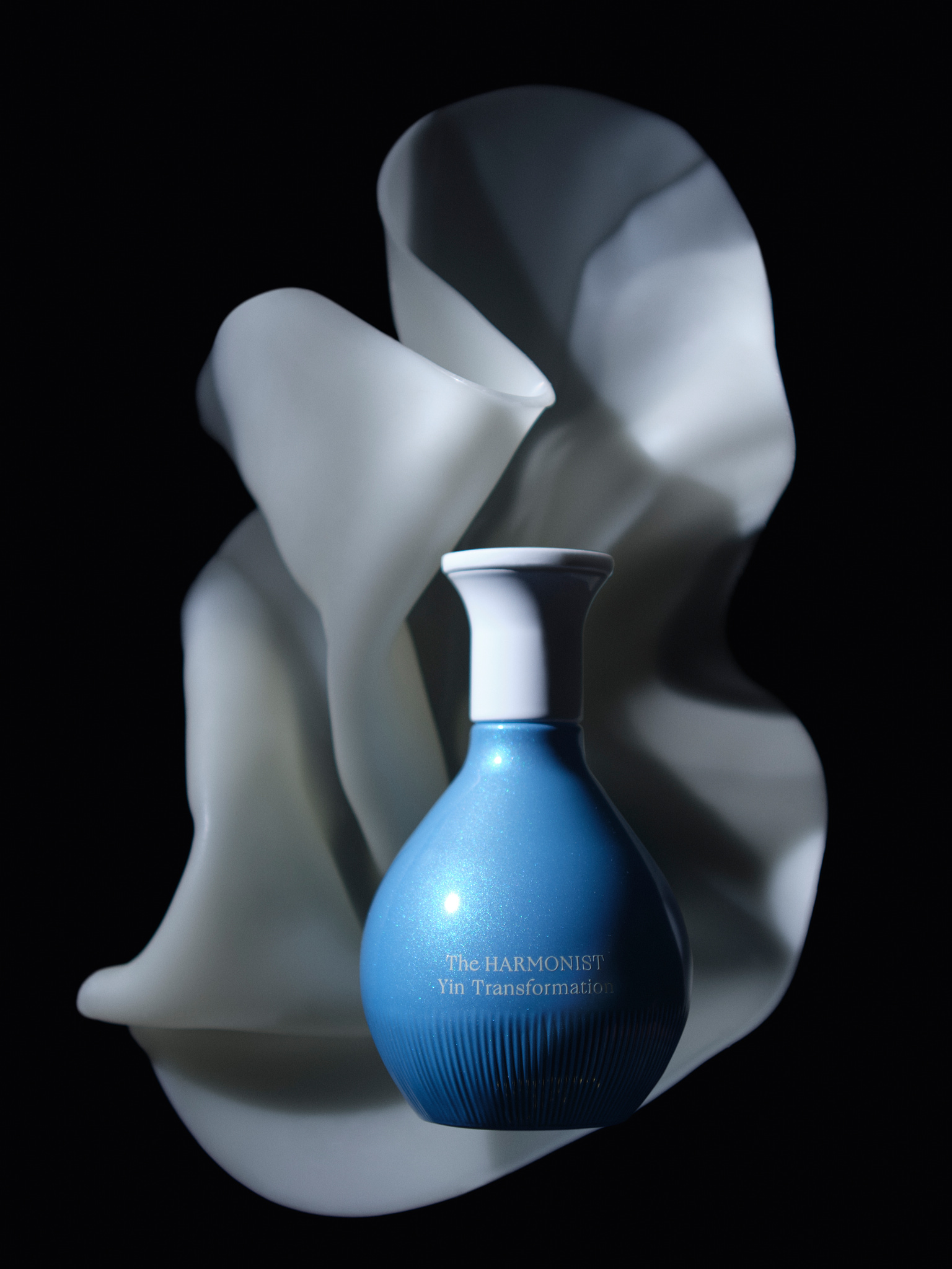 Blue bottle of The Harmonist Yin Transformation with white cap in front of flowing white shape.